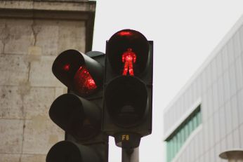 Eine rote Fußgängerampel zeigt ein stehendes Männchen, das Signal „Warten“. Im Hintergrund sind Gebäudefassaden zu sehen.
