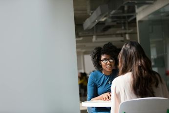 Das Bild zeigt zwei Frauen, die an einem Tisch in einem modernen, hellen Raum sitzen. Die Frau auf der linken Seite trägt eine Brille und ein blaues Oberteil, während sie der Frau gegenüber aufmerksam zuhört. Im Hintergrund sind weitere Personen und Büroeinrichtungen schemenhaft zu erkennen.