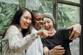 Drei lachende Personen stehen dicht beieinander und machen ein Selfie. Eine Person hält ein Smartphone in der Hand, während die anderen darauf zeigen. Im Hintergrund sind große Fenster und grüne Pflanzen zu sehen, was auf eine helle, natürliche Umgebung hindeutet.