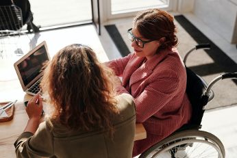 Frau sitzt im Rollstuhl am Tisch und spricht mit anderen Frau