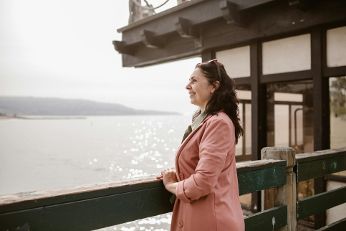 Das Bild zeigt eine Frau im pinken Blazer. Sie steht am Wasser und schaut in die Ferne