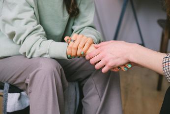 Das Bild zeigt zwei Personen, die Hände halten. Die Person links trägt eine hellgrüne Sweatshirtjacke und beige Hosen, während bei der Person rechts einen gemusterten Stoffärmel zu sehen ist.