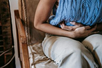 Das Bild zeigt eine Person, die auf einem gepolsterten Holzstuhl sitzt. Sie trägt eine beige Hose und hält ein blaues, gestricktes Kissen mit beiden Armen eng an ihren Bauch gedrückt.