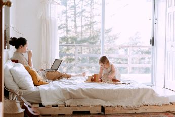 Frau sitzt mit Laptop gemeinsam mit ihrem Kind auf dem Bett