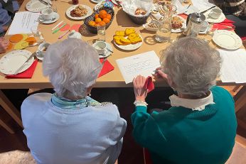 Eine Gruppe älterer Menschen sitzt an einem Tisch mit Kaffee, Tee, Gebäck und Obst. Zwei Frauen im Vordergrund schauen auf ein Blatt Papier mit einem Text. Auf dem Tisch liegen bunte Zettel und Stifte
