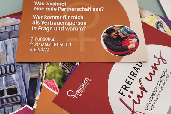 Das Bild zeigt ein Set aus Karten mit Themen für Frauen in der Rente. Eine Karte stellt Fragen zur Partnerschaft und Vertrauenspersonen, begleitet von einer kleinen Rundabbildung eines älteren und jüngeren Menschen in einer herzlichen Umarmung. Begriffe wie "Fürsorge", "Zusammenhalten" und "Einsam" sind auf der Karte aufgeführt. Das Design ist in warmen Farbtönen gehalten, mit dem Titel „Freiraum für uns“ auf einer anderen sichtbaren Karte.