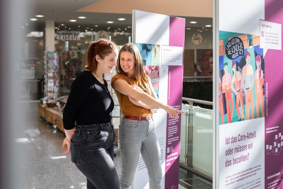 Zwei Frauen stehen nebeneinander in einem Einkaufszentrum und betrachten lächelnd eine Ausstellung mit Text- und Bildtafeln. Eine der Frauen zeigt auf eine der Tafeln. Die Ausstellung behandelt das Thema „Care-Arbeit“ und ist mit lila Elementen gestaltet.