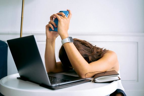 Eine Person sitzt an einem runden Tisch mit einem Laptop, einem Notizbuch und einem Smartphone. Sie hält das Smartphone mit beiden Händen hoch und lehnt ihren Kopf erschöpft auf die Arme.