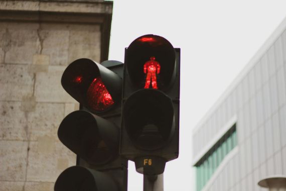 Eine rote Fußgängerampel zeigt ein stehendes Männchen, das Signal „Warten“. Im Hintergrund sind Gebäudefassaden zu sehen.
