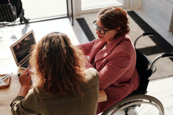 Frau sitzt im Rollstuhl am Tisch und spricht mit anderen Frau