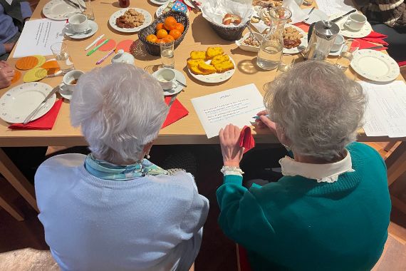 Eine Gruppe älterer Menschen sitzt an einem Tisch mit Kaffee, Tee, Gebäck und Obst. Zwei Frauen im Vordergrund schauen auf ein Blatt Papier mit einem Text. Auf dem Tisch liegen bunte Zettel und Stifte